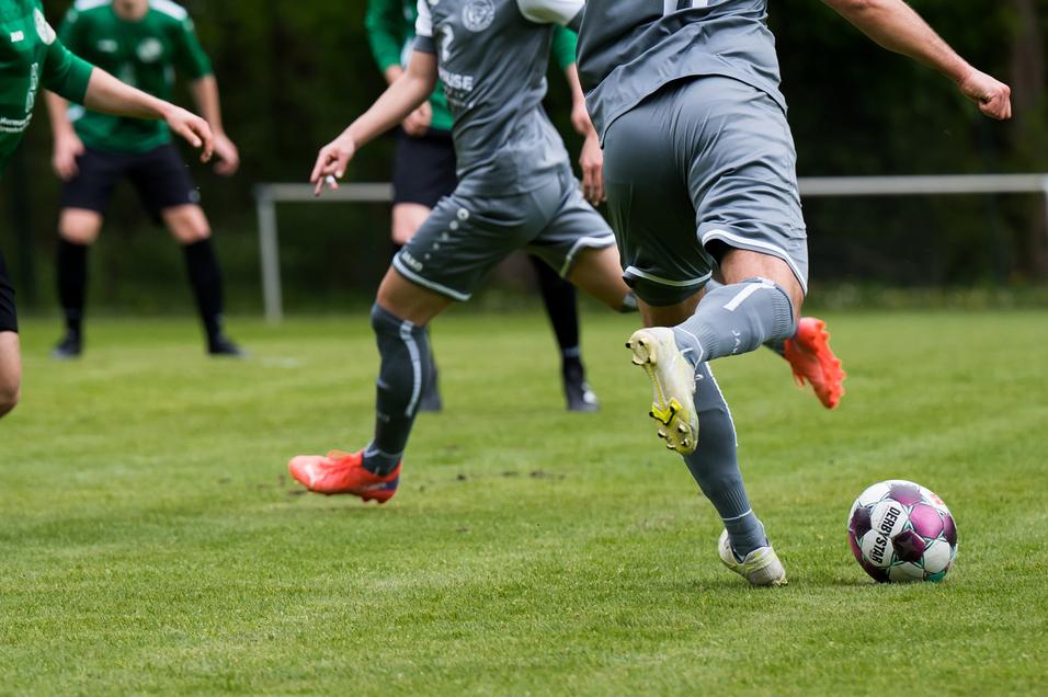 Drei Fußballspieler in grünen und grauen Trikots auf einem Rasenplatz, während ein Spieler den Ball zuspielt.