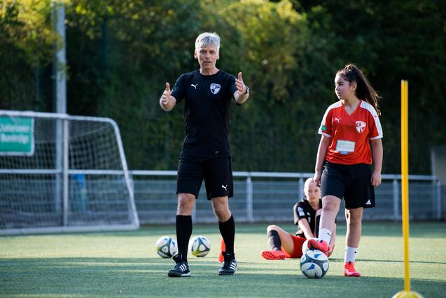 Trainer erklärt Technik beim Fußballtraining, während Spielerinnen auf dem Kunstrasen stehen und sitzen.