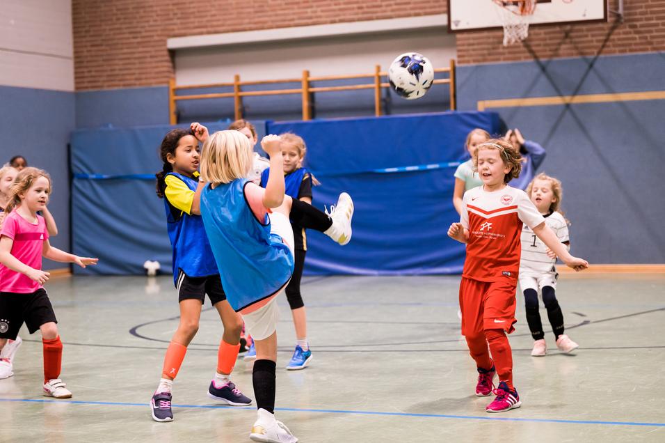Mädchen spielen Fußball in einer Halle, ein Mädchen hebt den Fuß zum Schuss, während andere zuschauen.