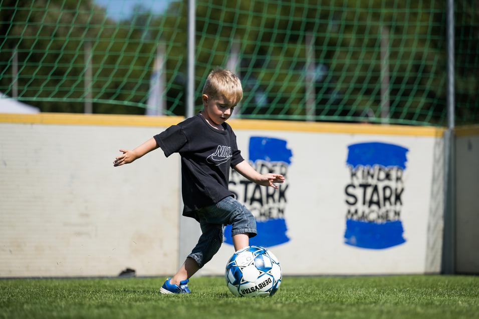 Kleiner Junge in schwarzem T-Shirt kickt einen Fußball auf einem grünen Rasenspielfeld. Im Hintergrund sind Wände sichtbar.