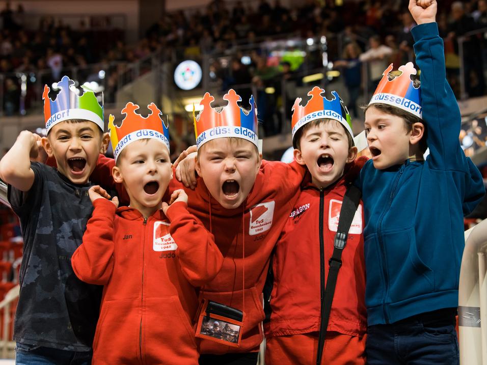 Gruppe von fünf jubelnden Kindern mit farbigen Kronen und roten Sweatshirts in einer Sportarena.