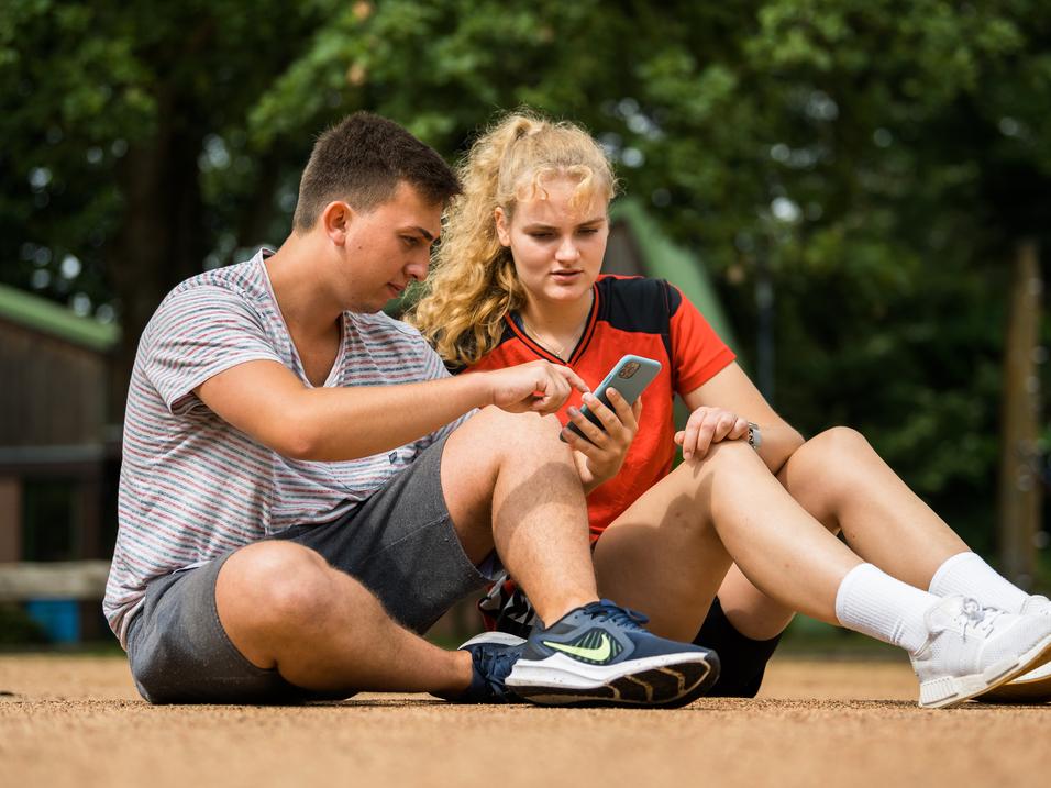 Zwei Jugendliche sitzen auf dem Boden und betrachten gemeinsam ein Smartphone. Ein Mädchen trägt ein rotes sportliches Oberteil.