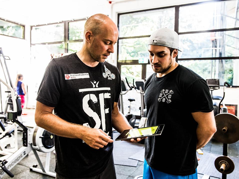Zwei Trainer im Fitnessstudio betrachten gemeinsam ein Tablet. Im Hintergrund sind Trainingsgeräte zu sehen.